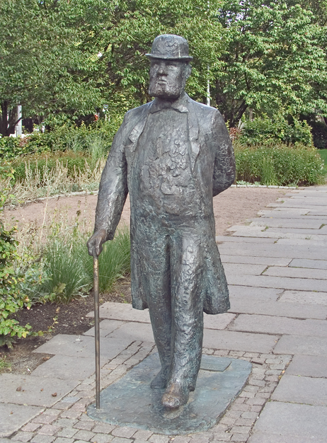 Staty av Charles Felix Lindberg. Han spatserar fram i hatt och käpp, med ena handen vilandes på ryggen.