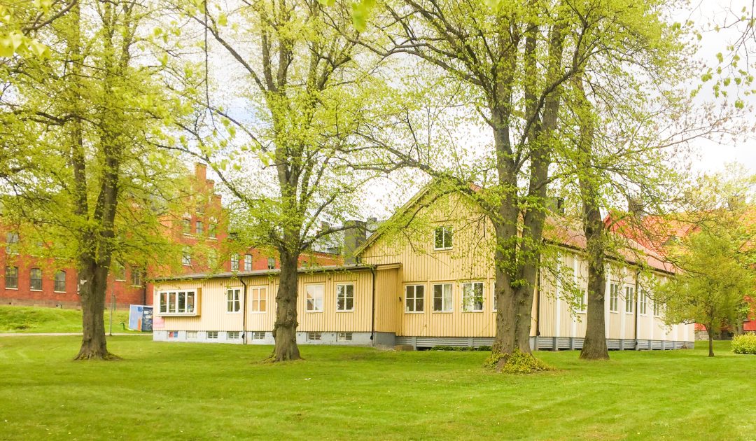 Ett gult trähus syns bakom några grönskande träd på en välklippt grön gräsmatta.