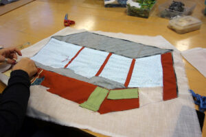 Ett broderi under arbete. Applikationer har lagts ut på tyget i formen av ett brunt hus med stora fönster.