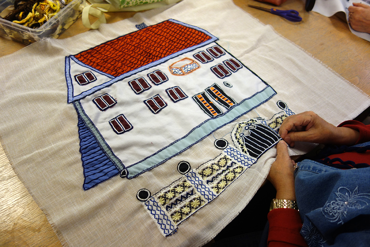Två händer arbetar med ett broderi som föreställer ett vitt hus med fönsterluckor, och med ett staket och en grind framför.