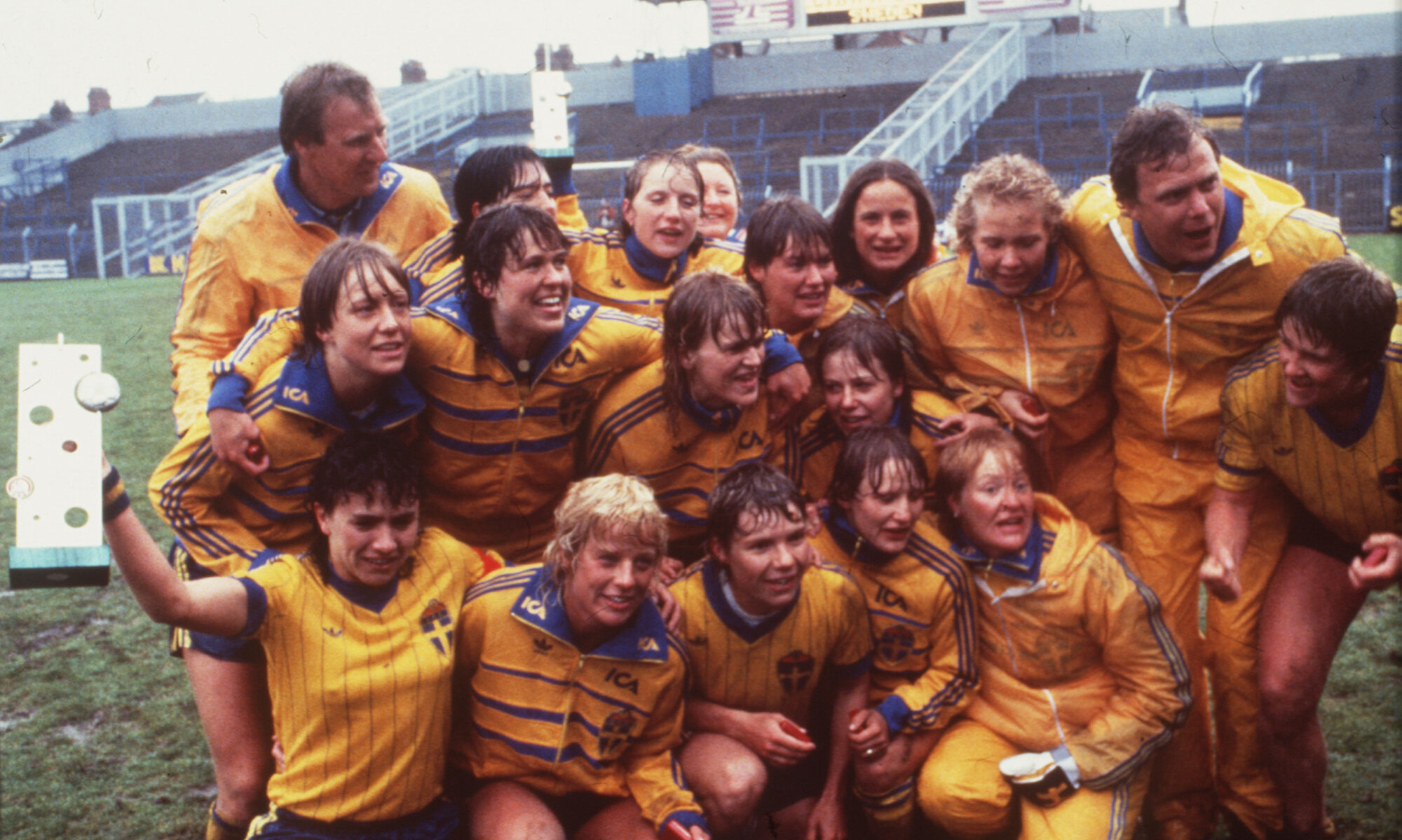 Landslaget i gula tröjor på en lerig fotbollsplan. De kramas och håller om varandra. En av spelarna håller i EM-pokalen..