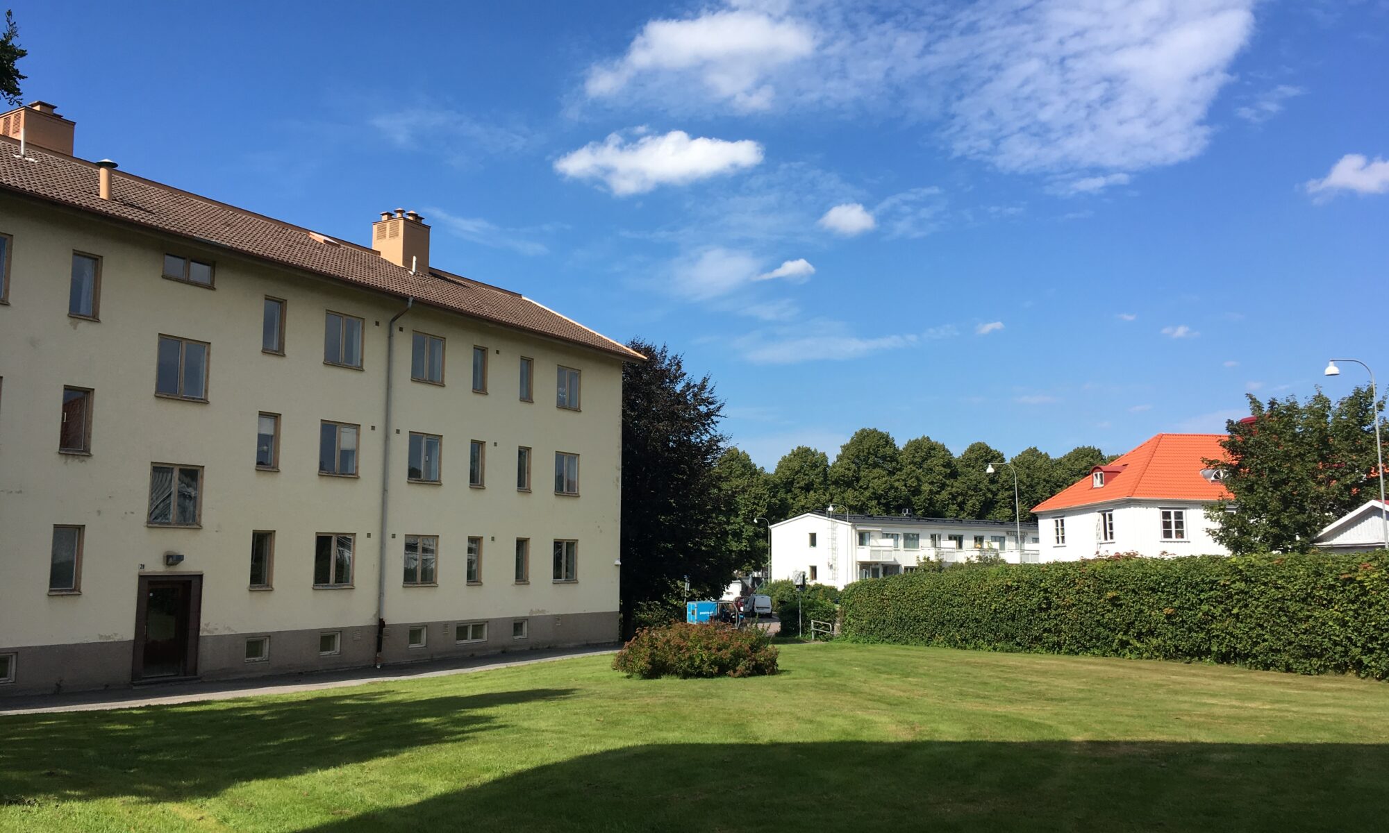 Stor grön gräsyta framför ett lägenhetshus i tre våningar. Solen skiner och det är sommar.