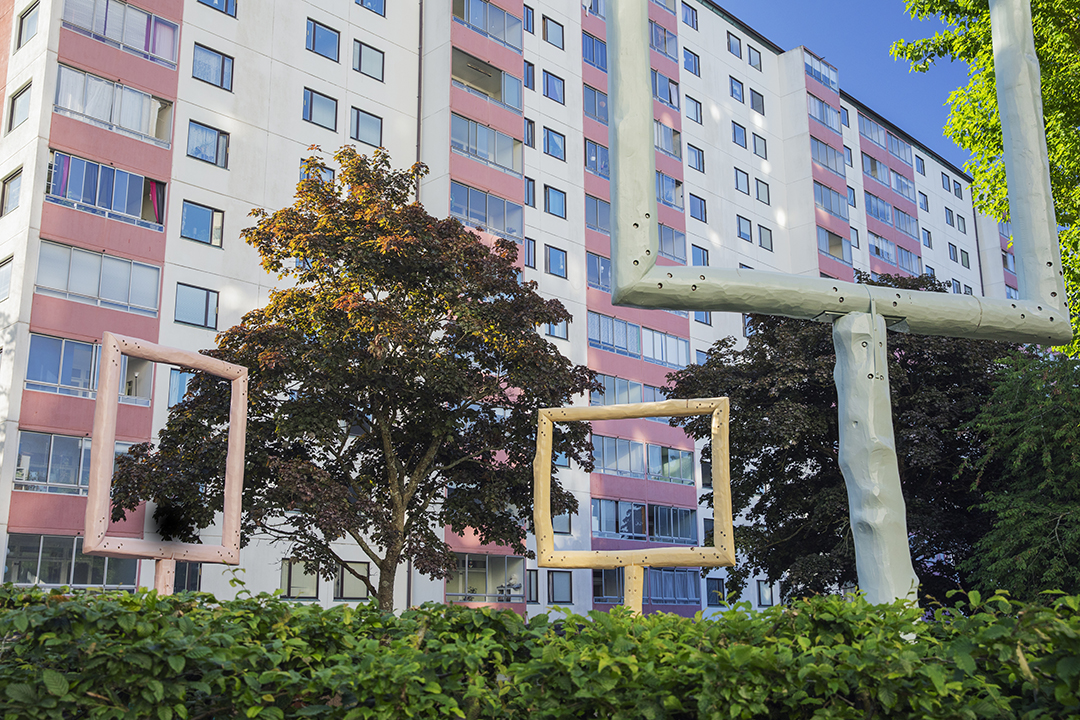 Tre fönsterskulpturer i rosa, gult och ljusgrön sticker upp bakom en häck framför höghusen i Kviberg.