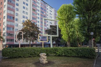 The Common Home En mindre träskulptur av en hög med böcker syns framför en häck. Bakom häcken tornar fönsterskulpturerna upp sig mot de höga husfasaderna.