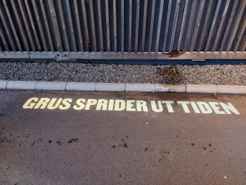 Grus sprider ut tiden Texten "Grus sprider ut tiden" projiceras på asfalten bredvid fasaden på ett parekringshus.